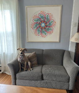 The spiraling whirl of Buddha Family is a very large framed print hung on a soft blue-hued wall, and a cute dog poses on a gray settee underneath.